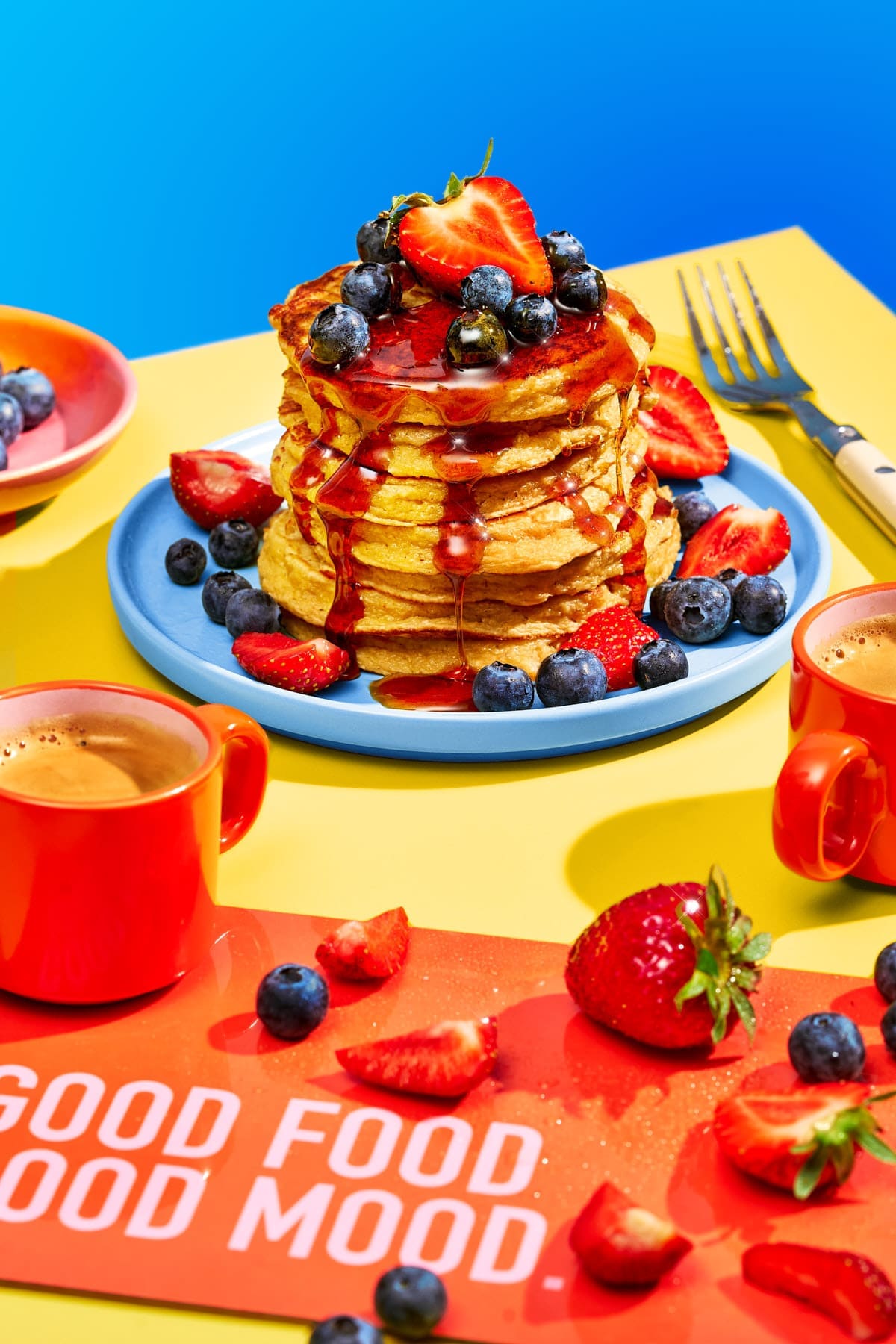 Hoher Stapel goldbrauner Protein Pancakes mit Ahornsirup, frischen Erdbeeren und Blaubeeren, serviert mit zwei Tassen Kaffee